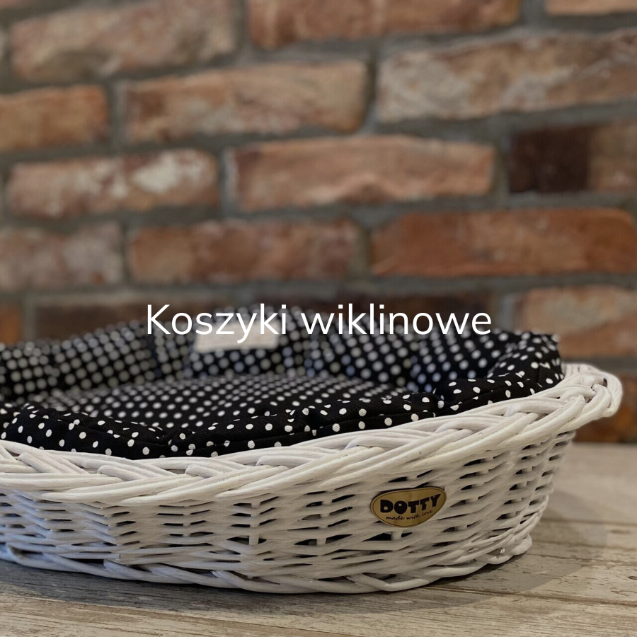 na tle ściany z cegieł na podłodze stoi koszyk wiklinowy malowany na biało z legowiskiem w środku , legowisko w kolorze czarnym w  białe groszki, widoczne złote logo DOTTY przyczepione do koszyczka